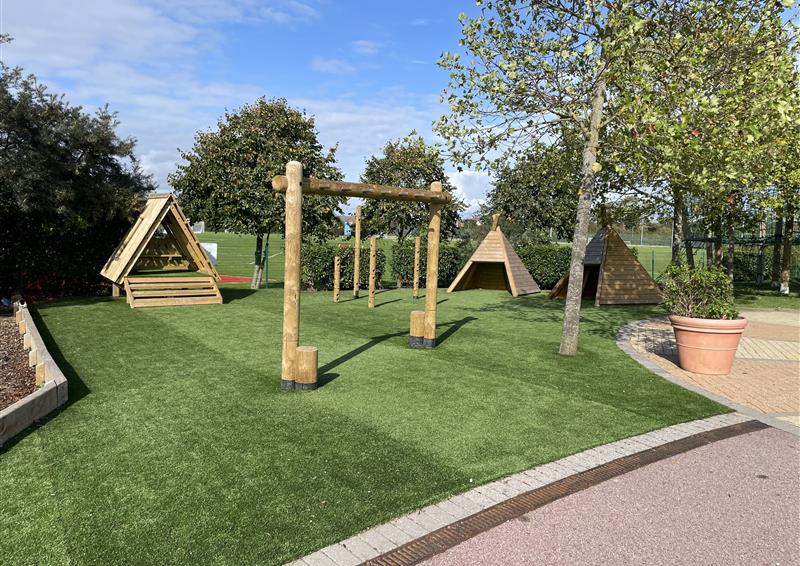 An artificial grass area with a variety of wooden play equipment installed on top of it. Surrounding the area is a beautiful resin bound path with trees and fields seen in the distance.